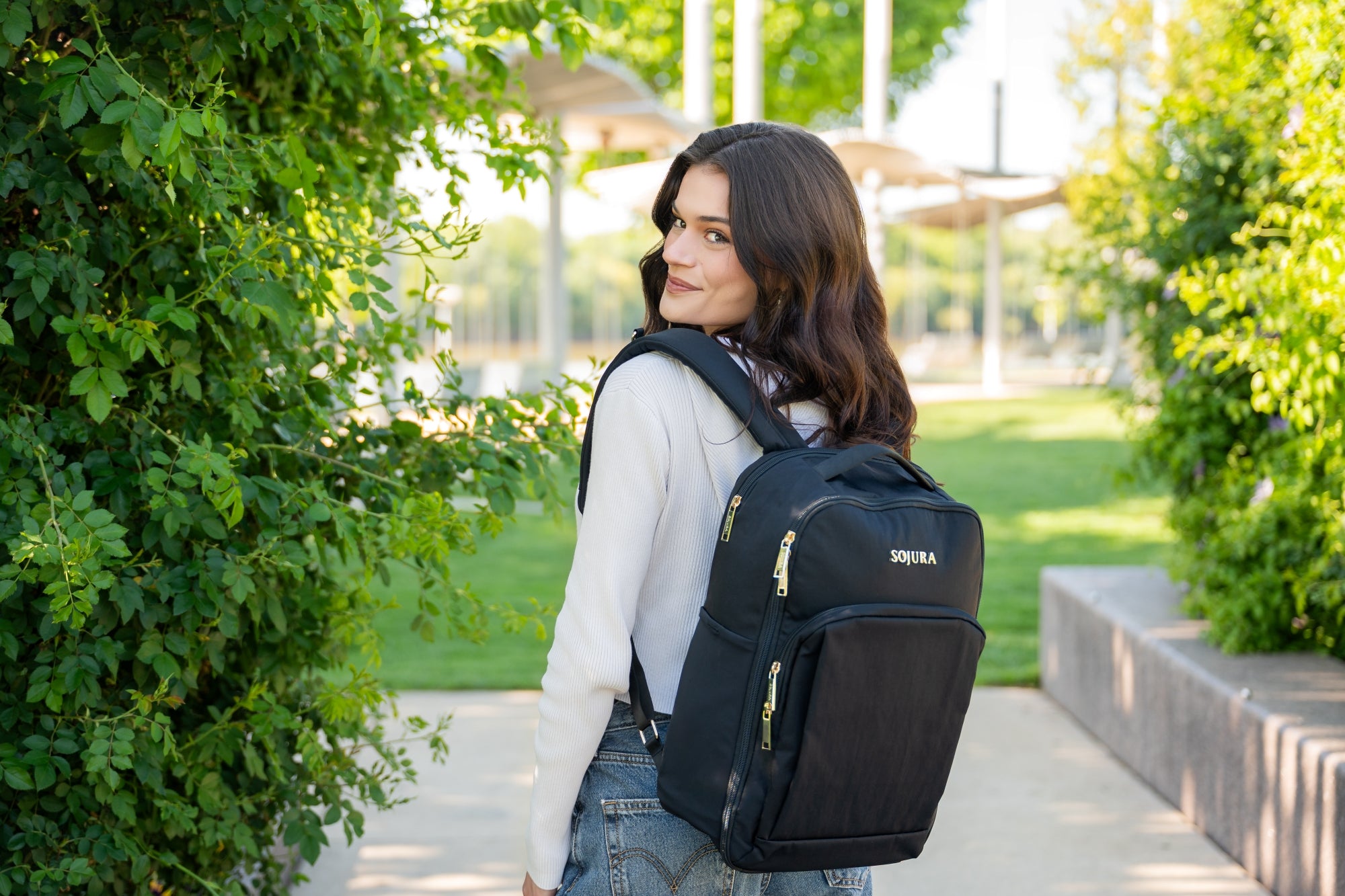 Woman wearing Sojura travel backpack looking over her shoulder while walking through greenery landscape